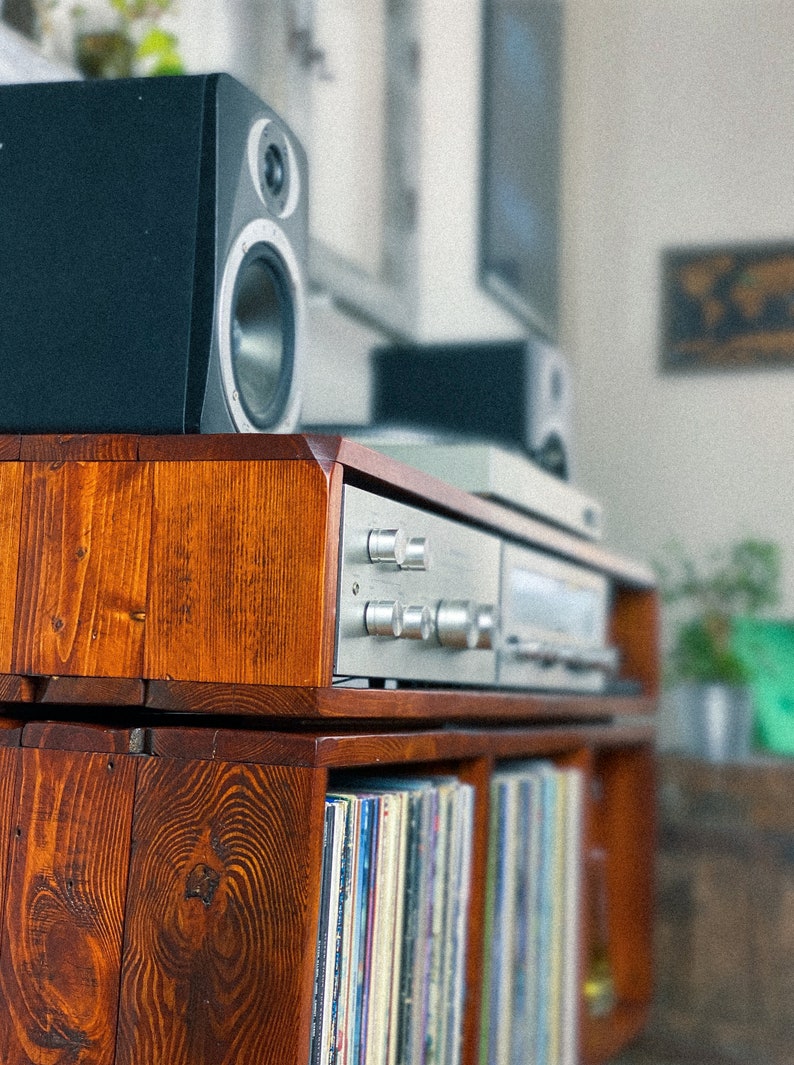 May include: A wooden record player stand with two shelves. The top shelf has a silver stereo receiver and two black speakers. The bottom shelf has a collection of vinyl records.
