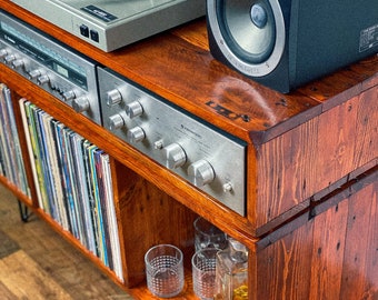 May include: A wooden record player stand with a turntable, amplifier, and speakers. The stand has shelves for storing records and a lower shelf for other items. The stand is made of reclaimed wood and has a rustic finish.