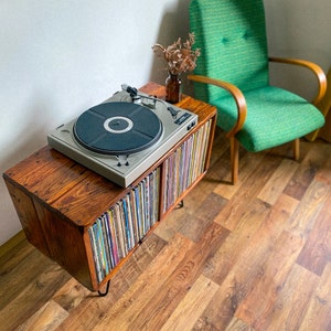 May include: A wooden record player stand with a turntable on top and shelves filled with vinyl records. The stand has black metal legs and is sitting on a wood floor.