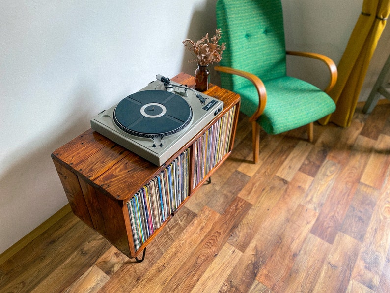 May include: A wooden record player stand with a turntable on top and shelves filled with vinyl records. The stand has black metal legs and is sitting on a wood floor.