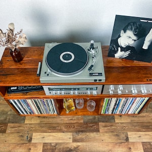 May include: A wooden record player stand with a turntable, amplifier, and shelves for vinyl records. The turntable is playing a record with the album cover "Heroes" by David Bowie visible on top of the stand.