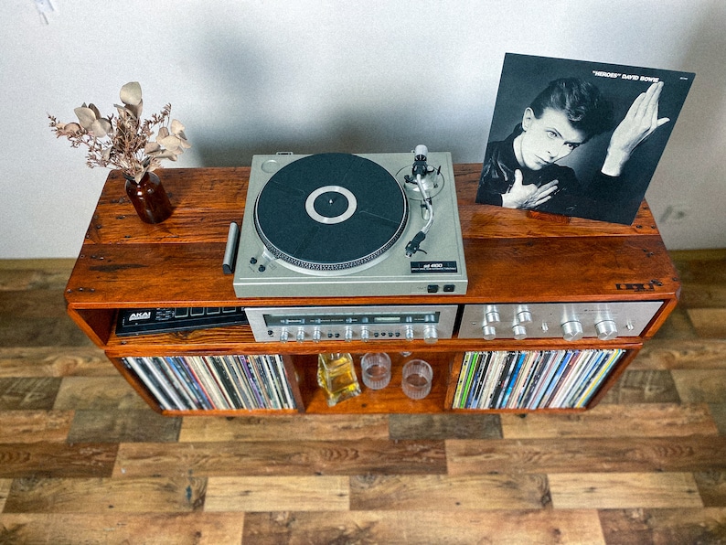 May include: A wooden record player stand with a turntable, amplifier, and shelves for vinyl records. The turntable is playing a record with the album cover "Heroes" by David Bowie visible on top of the stand.