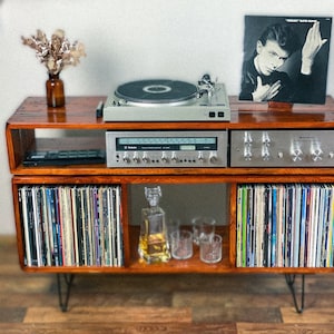 May include: A wooden record player stand with two shelves. The top shelf features a turntable, a stereo receiver, and a record album cover featuring David Bowie. The bottom shelf has a collection of vinyl records and a clear glass decanter with a stopper.