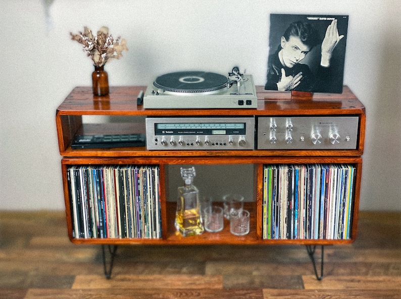 May include: A wooden record player stand with two shelves. The top shelf features a turntable, a stereo receiver, and a record album cover featuring David Bowie. The bottom shelf has a collection of vinyl records and a clear glass decanter with a stopper.
