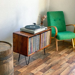 May include: A vintage record player sits on a wooden record cabinet with a collection of vinyl records. The cabinet has hairpin legs and is next to a green armchair.