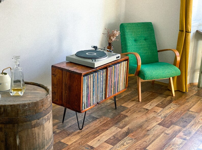 May include: A vintage record player sits on a wooden record cabinet with a collection of vinyl records. The cabinet has hairpin legs and is next to a green armchair.
