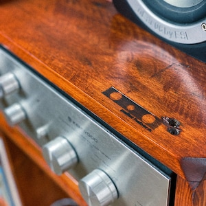 May include: A silver stereo receiver with three knobs and the text "7000" on the front. The receiver is sitting on a wooden shelf with a speaker on top.