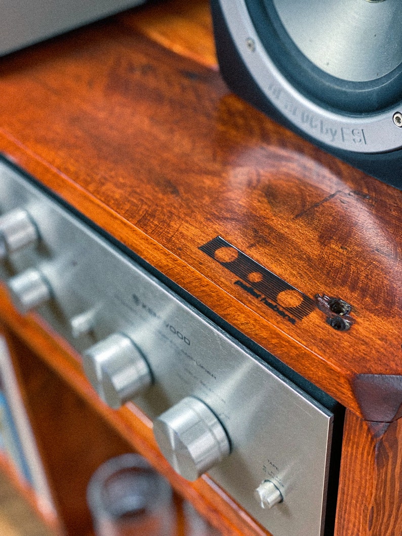 May include: A silver stereo receiver with three knobs and the text "7000" on the front. The receiver is sitting on a wooden shelf with a speaker on top.