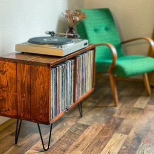 May include: A wooden record player stand with two shelves for vinyl records. The stand has black hairpin legs and is stained a dark brown color. A turntable sits on top of the stand.