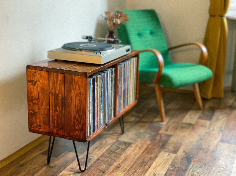 May include: A wooden record player stand with two shelves for vinyl records. The stand has black hairpin legs and is stained a dark brown color. A turntable sits on top of the stand.