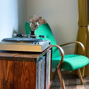 May include: A wooden record player stand with a turntable on top and a shelf for vinyl records. The stand is next to a green armchair with wooden arms.