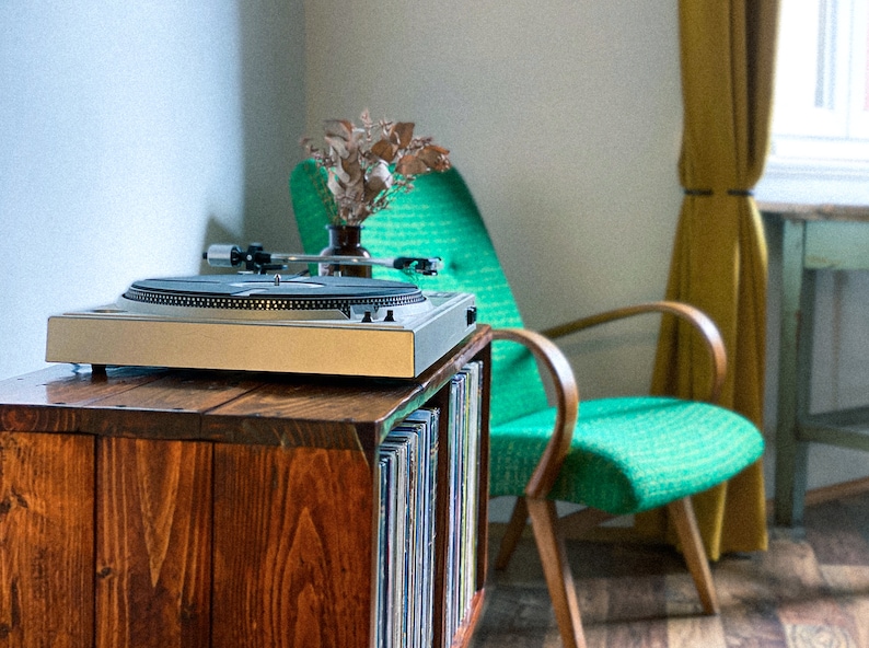 May include: A wooden record player stand with a turntable on top and a shelf for vinyl records. The stand is next to a green armchair with wooden arms.