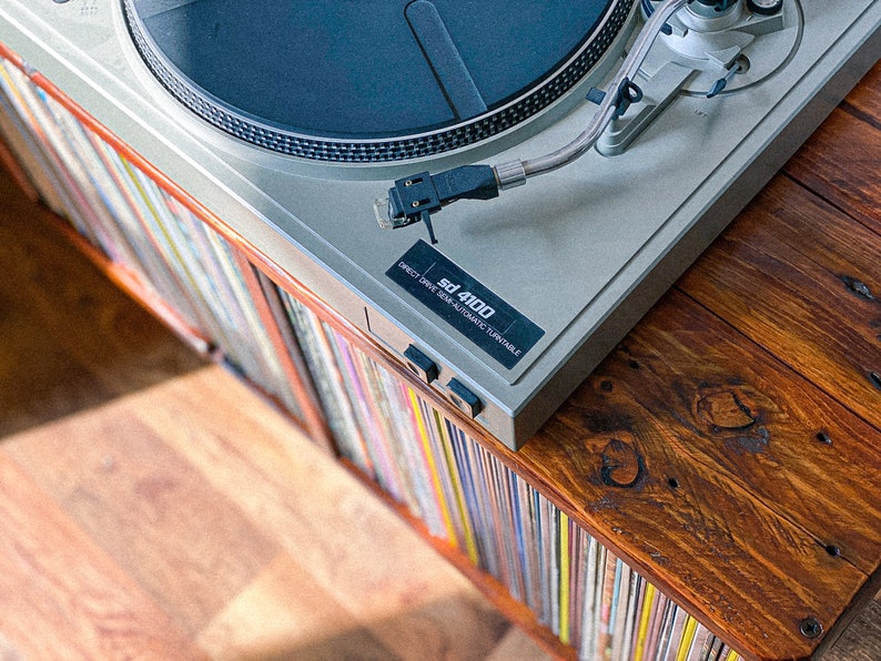 May include: A silver turntable with a black platter and a silver tonearm. The turntable is labeled "sd 4100 Direct Drive Semi-Automatic Turntable". The turntable is sitting on a wooden shelf with a collection of vinyl records.