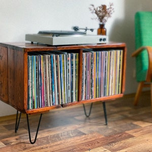 May include: A wooden record player stand with black hairpin legs. The stand has two shelves, each filled with vinyl records. A turntable sits on top of the stand.