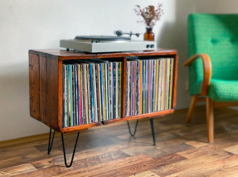 May include: A wooden record player stand with black hairpin legs. The stand has two shelves, each filled with vinyl records. A turntable sits on top of the stand.