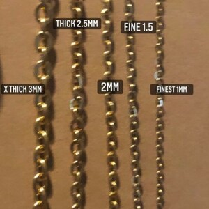 K&ouml;nnte beinhalten: F&uuml;nf goldene Ketten in verschiedenen St&auml;rken, beschriftet mit ihren Ma&szlig;en in Millimetern. Die Ketten sind von der dicksten zur d&uuml;nnsten angeordnet, wobei die dickste Kette mit "X THICK 3MM" beschriftet ist, gefolgt von "THICK 2.5MM", "2MM", "FINE 1.5" und "FINEST 1MM".