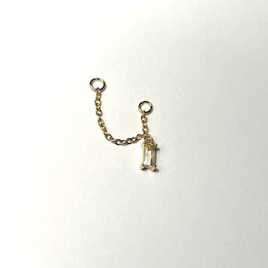 May include: A gold-colored double piercing earring with a chain and a rectangular, clear gemstone. The earring has two circular loops for piercing and a delicate chain connecting them. The gemstone is set in a gold-colored bezel.