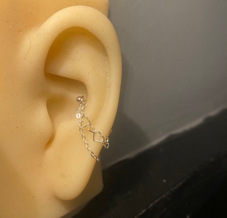 Op de afbeelding: Een zilveren oorbel met een kleine kogelknop en een delicate ketting. De ketting is versierd met drie hartvormige bedels. De oorbel wordt getoond op een mannequin-oor.