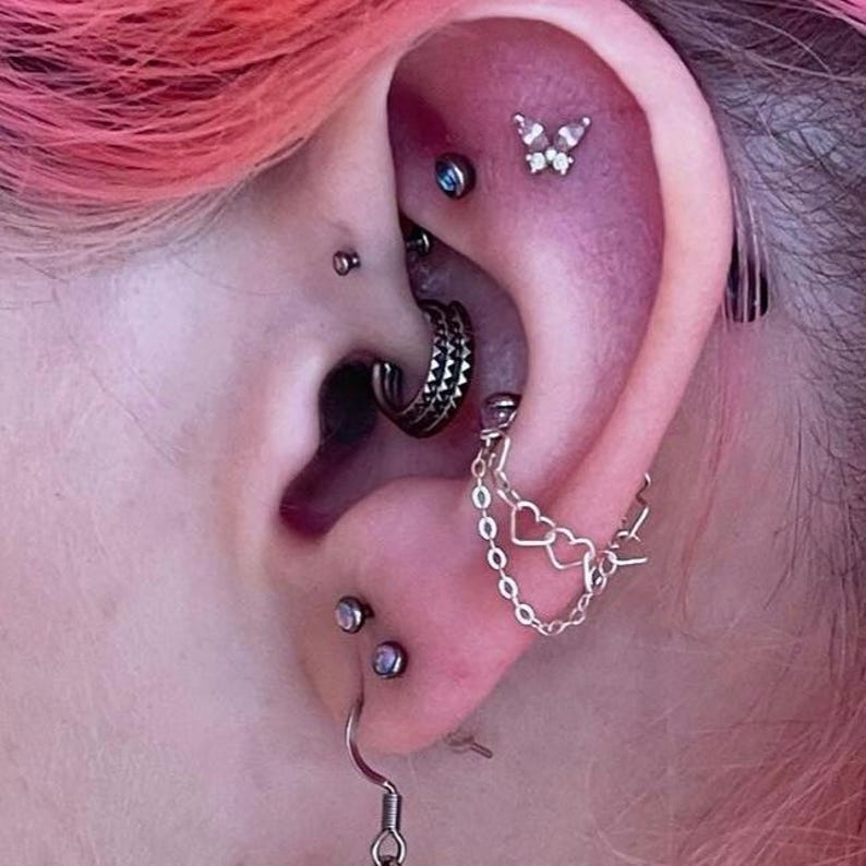 Op de afbeelding: Close-up van een oor met meerdere piercings. Het oor heeft verschillende sieraden, waaronder een vlinderstud, een ketting met hartjesbedels en verschillende oorringen en oorknopjes. De huid rond het oor is roze.