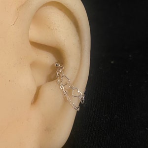 Op de afbeelding: Een zilveren oorbel met een ketting en twee hartvormige bedels. De oorbel is aan het oor bevestigd met een kleine kogelstud. Het oor is lichtbeige van kleur en de achtergrond is zwart.