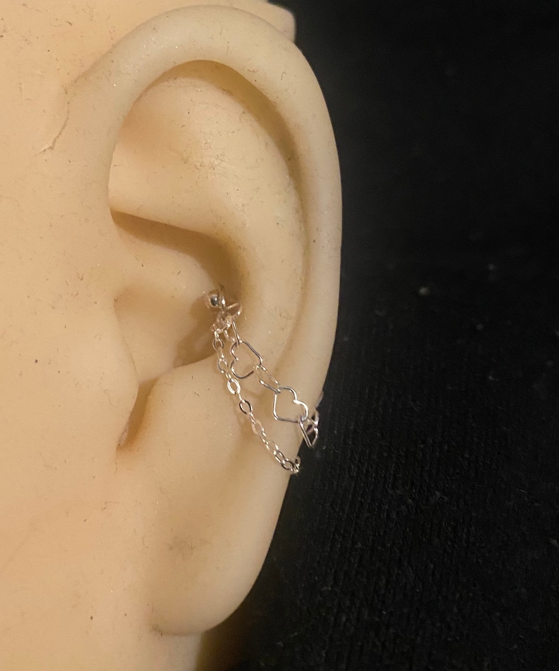 Op de afbeelding: Een zilveren oorbel met een ketting en twee hartvormige bedels. De oorbel is aan het oor bevestigd met een kleine kogelstud. Het oor is lichtbeige van kleur en de achtergrond is zwart.