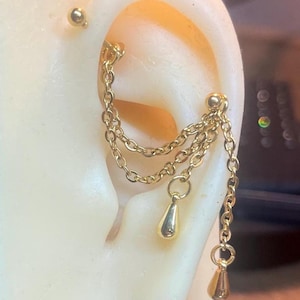 May include: Gold-coloured ear jewellery featuring a small stud and a chain design. The chain dangles with teardrop-shaped charms. The jewellery is displayed on a light-coloured ear model, showcasing the design and details.