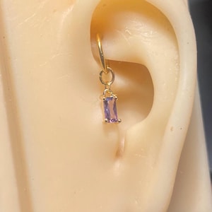 May include: A gold-colored hoop earring with a small, dangling rectangular purple gemstone. The earring is shown on a mannequin ear, highlighting the jewelry's design and color.