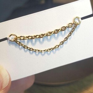 Op de afbeelding: Een delicate gouden kettingoorbel met twee strengen schakelketting. De oorbel is bevestigd aan een kleine ring aan de bovenkant en wordt gepresenteerd op een witte kaart. De ketting is ongeveer 5 cm lang.