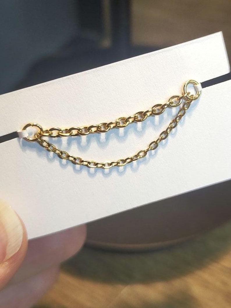 Op de afbeelding: Een delicate gouden kettingoorbel met twee strengen schakelketting. De oorbel is bevestigd aan een kleine ring aan de bovenkant en wordt gepresenteerd op een witte kaart. De ketting is ongeveer 5 cm lang.