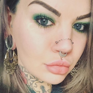 May include: Close-up of a person's face with green eyeshadow and blue eyes. The person has multiple facial piercings, including a septum ring, a nose chain, and a lip piercing. Large, ornate earrings are visible.