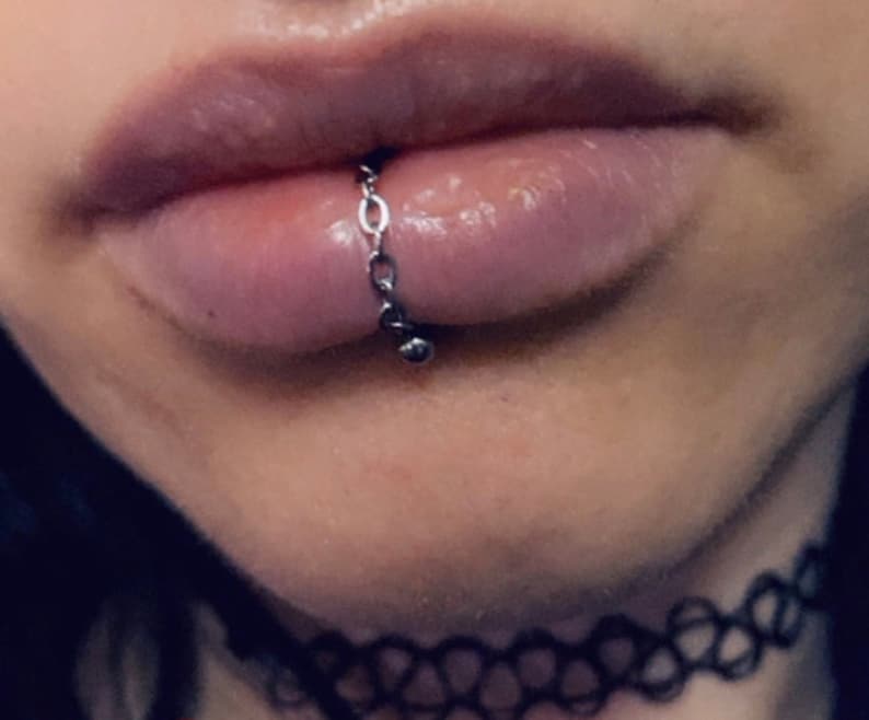 K&ouml;nnte beinhalten: Nahaufnahme einer Unterlippe mit einem silbernen Kettenpiercing. Die Kette ist an beiden Enden an einer kleinen silbernen Kugel befestigt.