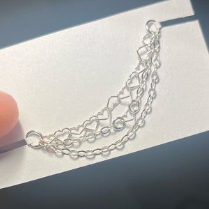 Op de afbeelding: Zilveren oormanchet met drie fijne kettingen. De bovenste ketting heeft een geometrisch ontwerp met een vierkant en een hart. De middelste ketting heeft hartvormige schakels en de onderste ketting is een klassiek kettingontwerp.