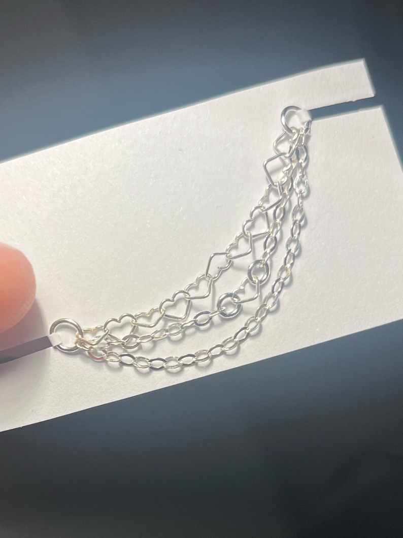 Op de afbeelding: Zilveren oormanchet met drie fijne kettingen. De bovenste ketting heeft een geometrisch ontwerp met een vierkant en een hart. De middelste ketting heeft hartvormige schakels en de onderste ketting is een klassiek kettingontwerp.