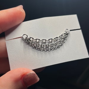 Op de afbeelding: Zilverkleurig ketting-sieraad met drie parallelle strengen, gehouden tegen een witte kaart. De kettingen zijn gemaakt van kleine, in elkaar grijpende schakels. Het sieraad wordt gepresenteerd op een witte kaart met een zwarte achtergrond.