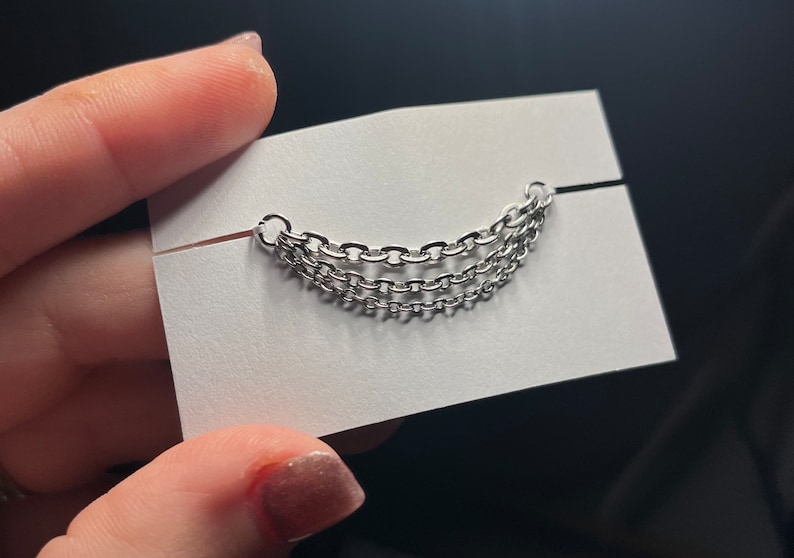 Op de afbeelding: Zilverkleurig ketting-sieraad met drie parallelle strengen, gehouden tegen een witte kaart. De kettingen zijn gemaakt van kleine, in elkaar grijpende schakels. Het sieraad wordt gepresenteerd op een witte kaart met een zwarte achtergrond.
