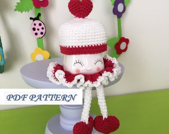 May include: White crocheted mug cozy with red heart accents and a smiling face. The cozy has white crocheted legs with red heart feet. The cozy is sitting on a purple pedestal. The text "PDF PATTERN" is visible in the image.
