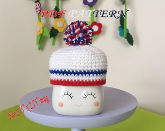 May include: A white ceramic mug with a smiling face and pink cheeks, topped with a crocheted white hat featuring blue and red stripes and a multi-colored pom-pom. The text PDF PATTERN and For 2.75" x 2.75" mug are visible on the image.