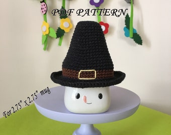 May include: A black crocheted pilgrim hat with a brown band and a yellow buckle. The hat is sitting on top of a white mug with a smiley face. The mug is on a purple cake stand. The text "PDF PATTERN" is visible in the image. The text "For 2.75" x 2.75" mug" is also visible in the image.