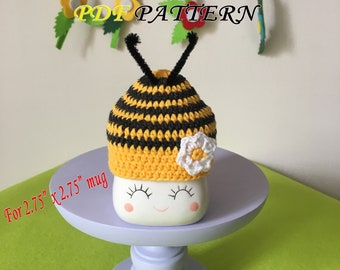 May include: A crocheted bee mug cozy in yellow and black stripes with a white flower accent. The cozy is designed to fit a 2.75 inch by 2.75 inch mug.  The text "PDF PATTERN" is visible in the image.