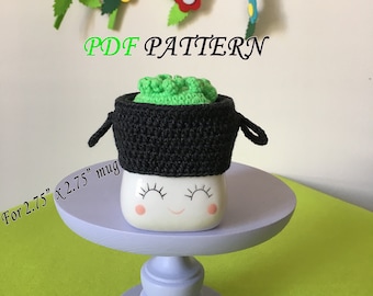 May include: A crochet mug cozy in black and green, with a green top and black bottom. The cozy is designed to fit a 2.75" x 2.75" mug. The text "PDF PATTERN" is written in white letters on a black background. The cozy is sitting on a white mug with a happy face.