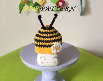 May include: A crochet bee hat with yellow and black horizontal stripes, two black pipe cleaner antennae, and a white flower applique. The hat is displayed on a white ceramic base with a printed face. The text PDF PATTERN is visible above.