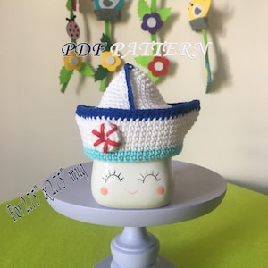 May include: White crocheted sailor hat with blue trim and a red and white life preserver design. The hat is sitting on a white mug with a smiley face. The text "For 2.75" x 2.75" mug" is written below the mug.