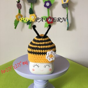 May include: A crocheted bee mug cozy in yellow and black stripes with a white flower accent. The cozy is designed to fit a 2.75 inch by 2.75 inch mug.  The text "PDF PATTERN" is visible in the image.