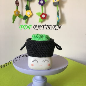 Pode incluir: Um aconchegante de caneca de crochê em preto e verde, com a parte superior verde e a inferior preta. O aconchegante é projetado para caber em uma caneca de 2,75" x 2,75". O texto "PDF PATTERN" está escrito em letras brancas sobre fundo preto. O aconchegante está sentado em uma caneca branca com um rosto sorridente.
