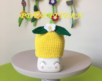 May include: A yellow crocheted lemon-shaped hat with two dark green leaves and a small white flower with a yellow center on top. The hat sits on a white ceramic mug featuring a printed face with closed eyes, long eyelashes, and pink cheeks.