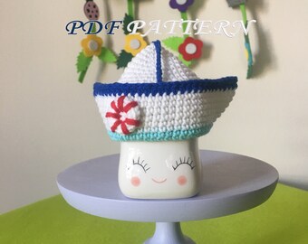 May include: A white ceramic mug with a painted smiling face wearing a white, blue, and light blue crocheted sailor hat featuring a red life preserver accent. The text PDF PATTERN is visible above the mug. The mug sits on a light purple stand.