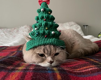Puede incluir: Un gato de pelo corto con un gorro de árbol de Navidad de crochet verde con adornos rojos y blancos. El gato está tumbado en una manta de cuadros rojos y negros.