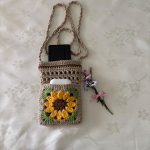 Op de afbeelding: Een beige gehaakte crossbodytas met een zonnebloemontwerp op de voorzak. De zonnebloem heeft gele bloemblaadjes, een bruin hart en groene bladeren. De tas heeft een lange riem en bevat een zwarte tablet en witte oortelefoons.