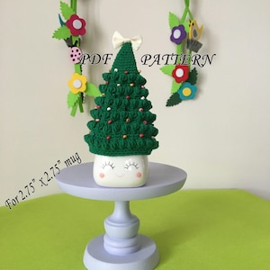 May include: Crocheted Christmas tree mug cozy in green with white beads and a white bow. The cozy fits a 2.75 inch by 2.75 inch mug. The text "PDF PATTERN" is visible in the image.