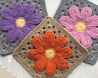 Op de afbeelding: Close-up van vier gehaakte vierkanten met 3D bloemmotieven. De bloemen zijn in tinten paars, roze en oranje, met gele centra. De vierkanten zijn grijs en bruin van kleur. Het woord "3D" staat in de linker benedenhoek.